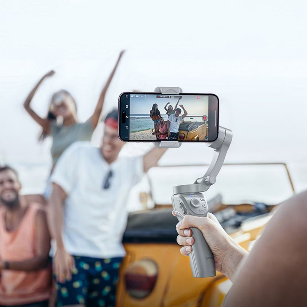 Person holding a smartphone gimbal with a video of people on a beach displayed, with a blurred background of people on a boat.