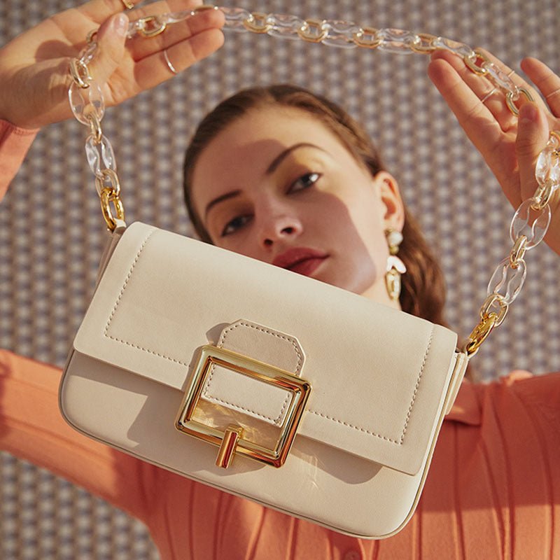 Woman holding a beige handbag with a gold clasp and clear chain strap. Emriavaly
